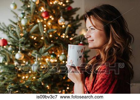 Stock Photo - Woman drinking coffee beside decorated Christmas tree. Fotosearch