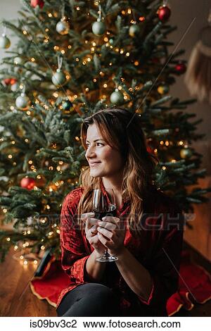Stock Image - Woman drinking wine beside decorated Christmas tree. Fotosearch