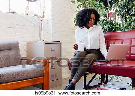 Stock Image - Woman in office sitting on sofa using laptop. Fotosearch