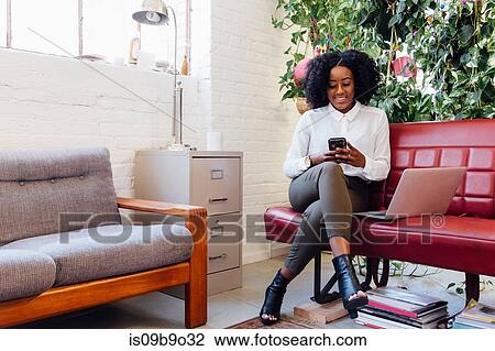 Stock Image - Woman in office sitting on sofa with laptop, texting on smartphone. Fotosearch