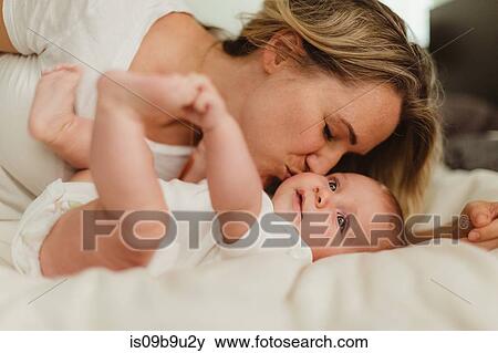 Stock Photo - Woman kissing baby daughter lying on bed. Fotosearch