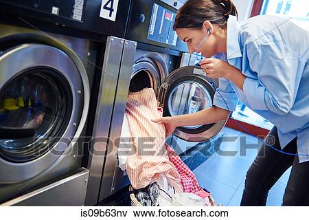 Woman listening to headphones inserting laundry into washing machine at laundrette View Large Photo Image Stock Photo - Woman listening to headphones inserting laundry into washing machine at laundrette. Fotosearch
