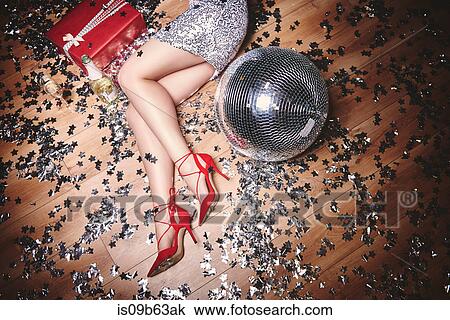 Stock Image - Woman lying on floor at party, surrounded by glitter, champagne bottle and disco ball, overhead view. Fotosearch