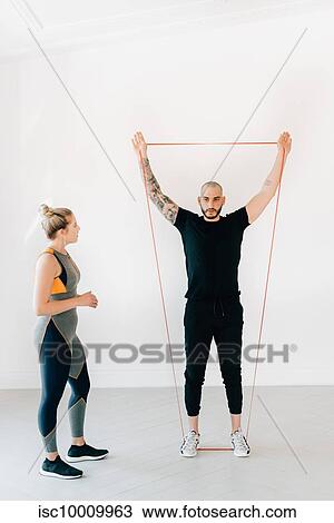 Stock Image - Woman observing fitness instructor using resistance band in studio. Fotosearch