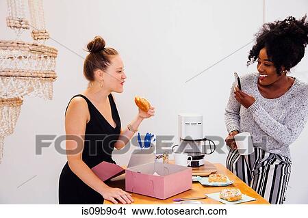 Stock Photograph - Woman photographing friend eating doughnut. Fotosearch