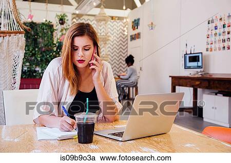 Stock Image - Woman sitting at desk in creative studio making telephone call on smartphone. Fotosearch