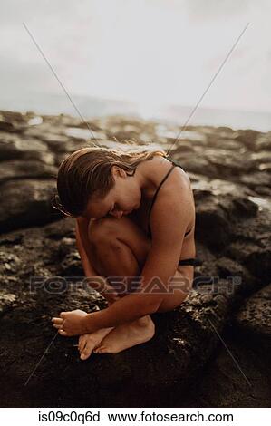 Stock Image - Woman sitting curled up on rocks by sea, Princeville, Hawaii, US. Fotosearch