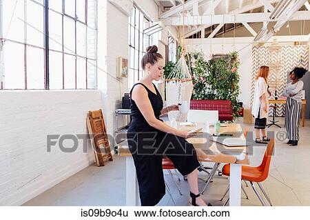 Stock Photograph - Woman sitting on desk in industrial office building texting on smartphone. Fotosearch