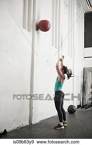 Stock Photo - Woman throwing fitness ball against wall in cross training gym. Fotosearch
