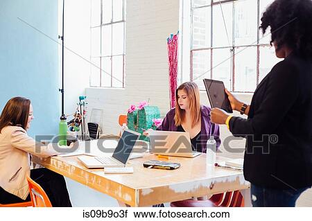 Stock Image - Woman with digital tablet speaking to colleagues working at desk. Fotosearch