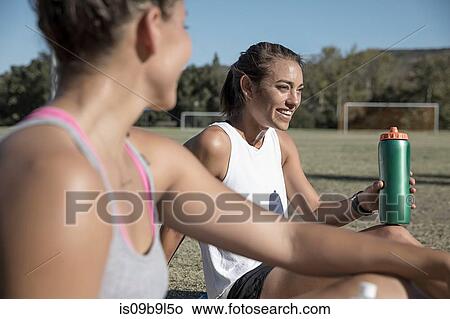 Picture - Women sitting on football pitch drinking from water bottle. Fotosearch