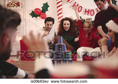 Stock Photo - Young adult friends throwing ball at paper cup pyramid at christmas party. Fotosearch