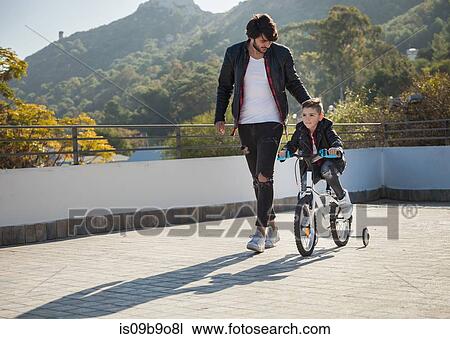 Young boy riding bike with stabilizers, father walking beside him View Large Photo Image Stock Image - Young boy riding bike with stabilizers, father walking beside him. Fotosearch