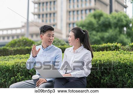 Stock Image - Young businesswoman and man using laptop and chatting on city seat, Shanghai, China. Fotosearch