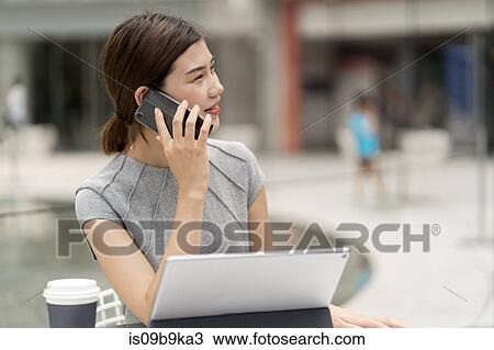 Stock Image - Young city businesswoman making smartphone call from sidewalk cafe. Fotosearch