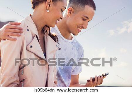 Picture - Young couple against blue sky looking at smartphone at roof party. Fotosearch