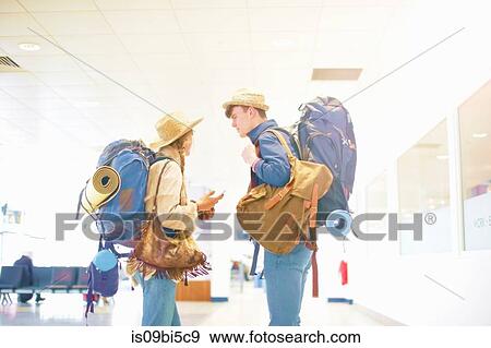 Stock Photo - Young couple at airport, carrying backpacks, standing face to face, talking. Fotosearch