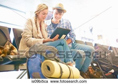 Stock Image - Young couple at airport, sitting, surrounded by backpacks, using digital tablet, low angle view. Fotosearch
