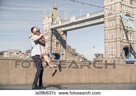 Stock Image - Young couple hugging outdoors, Tower Bridge in background, London, England, UK. Fotosearch