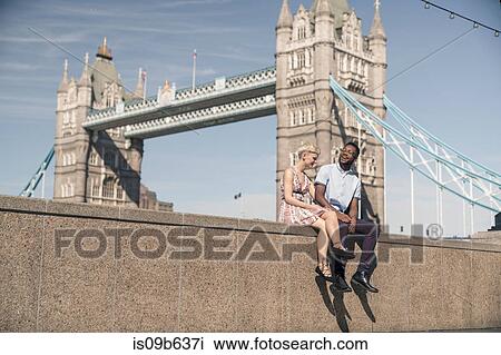 Stock Image - Young couple sitting on wall, smiling, Tower Bridge in background, London, England, UK. Fotosearch