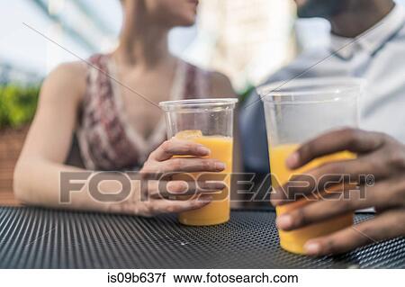 Stock Image - Young couple sitting outdoors, holding plastic drinking glasses, mid section. Fotosearch