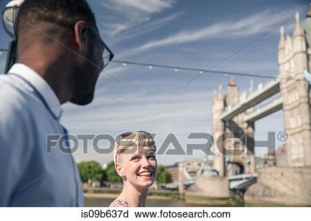 Stock Image - Young couple walking outdoors, smiling, Tower Bridge in background, London, England, UK. Fotosearch