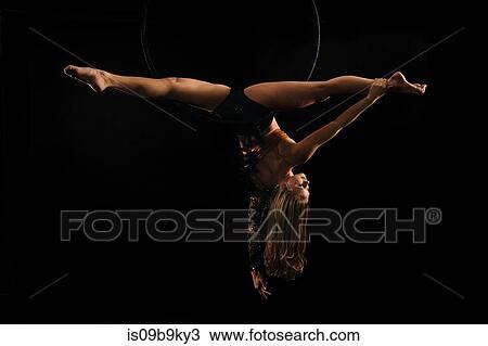 Stock Image - Young female aerial acrobat doing splits hanging upside down from hoop, black background. Fotosearch