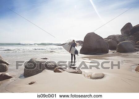 Stock Image - Young female surfer carrying surfboard looking out at ocean waves from beach, full length, Cape Town, Western Cape, South Africa. Fotosearch