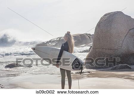 Stock Image - Young female surfer carrying surfboard looking out at ocean waves from beach, Cape Town, Western Cape, South Africa. Fotosearch