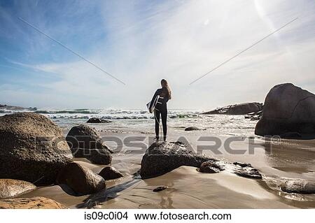 Picture - Young female surfer looking out from beach rock, rear view, Cape Town, Western Cape, South Africa. Fotosearch