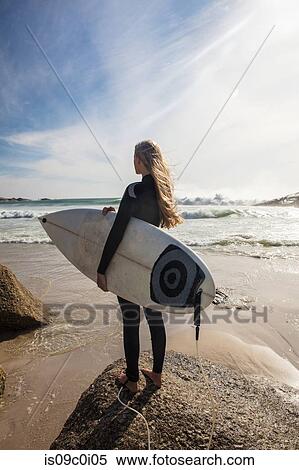 Stock Photography - Young female surfer looking out from beach, rear view, Cape Town, Western Cape, South Africa. Fotosearch