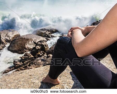 Stock Photography - Young female surfer sitting on beach rock amongst ocean waves, cropped, Cape Town, Western Cape, South Africa. Fotosearch