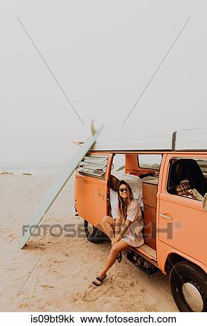 Stock Image - Young female surfer sitting on recreational vehicle on beach, portrait, Jalama, Ventura, California, USA. Fotosearch