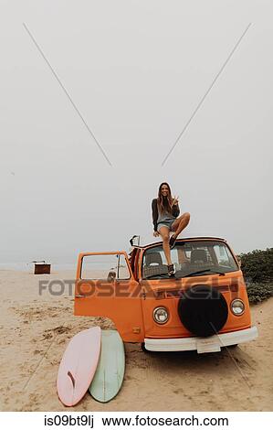 Stock Image - Young female surfer sitting on recreational vehicle roof at beach, portrait, Jalama, California, USA. Fotosearch