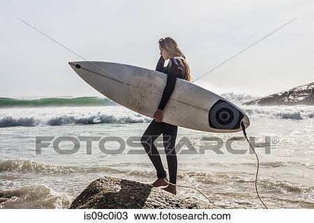 Stock Image - Young female surfer stepping onto beach rock, Cape Town, Western Cape, South Africa. Fotosearch
