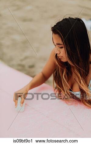 Stock Image - Young female surfer waxing surfboard at beach, Ventura, California, USA. Fotosearch