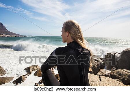 Stock Image - Young female surfer with long blond hair sitting on beach rock, rear view, Cape Town, Western Cape, South Africa. Fotosearch