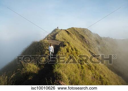 Stock Photograph - Young female tourist hiking on top of misty mountain, rear view, Bali, Indonesia. Fotosearch