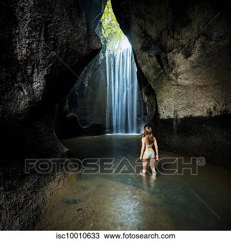 Stock Image - Young female tourist in bikini looking at rock formation waterfall, rear view, Bali, Indonesia. Fotosearch