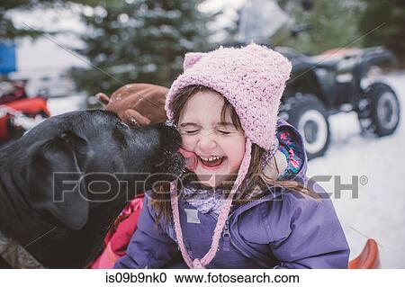 Stock Image - Young girl with dog in snowy landscape, dog licking girl's face. Fotosearch