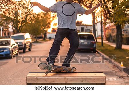 Stock Image - Young male skateboarder balancing on top of ramp on suburban street at sunset, neck down. Fotosearch