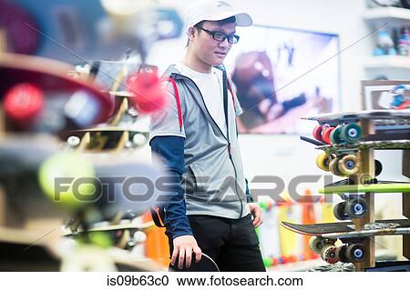 Stock Image - Young male skateboarder looking at skateboards in skateboard shop. Fotosearch