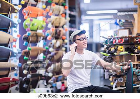 Stock Image - Young male skateboarder making smartphone call in skateboard shop. Fotosearch