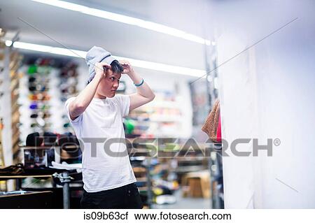 Stock Image - Young male skateboarder trying on knit hat in skateboard shop. Fotosearch