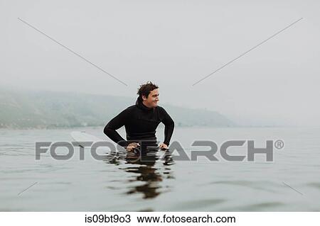 Stock Image - Young male surfer sitting on surfboard in calm misty sea, Ventura, California, USA. Fotosearch