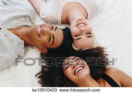Stock Image - Young man and two female friends lying on bed laughing, overhead view. Fotosearch