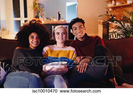 Stock Image - Young man and young female friends watching tv sitting on living room sofa with bowl of popcorn. Fotosearch