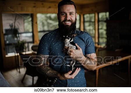 Stock Photo - Young man holding pet dog in living room, portrait. Fotosearch