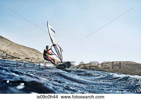 Picture - Young man leaning back windsurfing ocean waves, surface level view, Limnos, Khios, Greece. Fotosearch