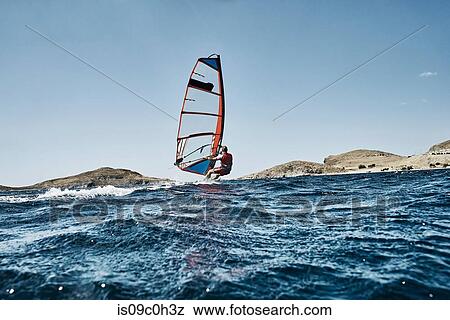 Stock Photo - Young man leaning back windsurfing ocean waves, side view, Limnos, Khios, Greece. Fotosearch